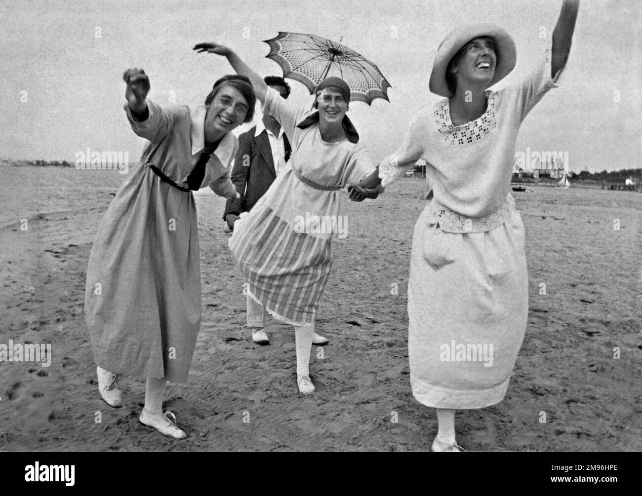 Three women striking funny poses on a beach.  A man in the background holds a parasol. Stock Photo