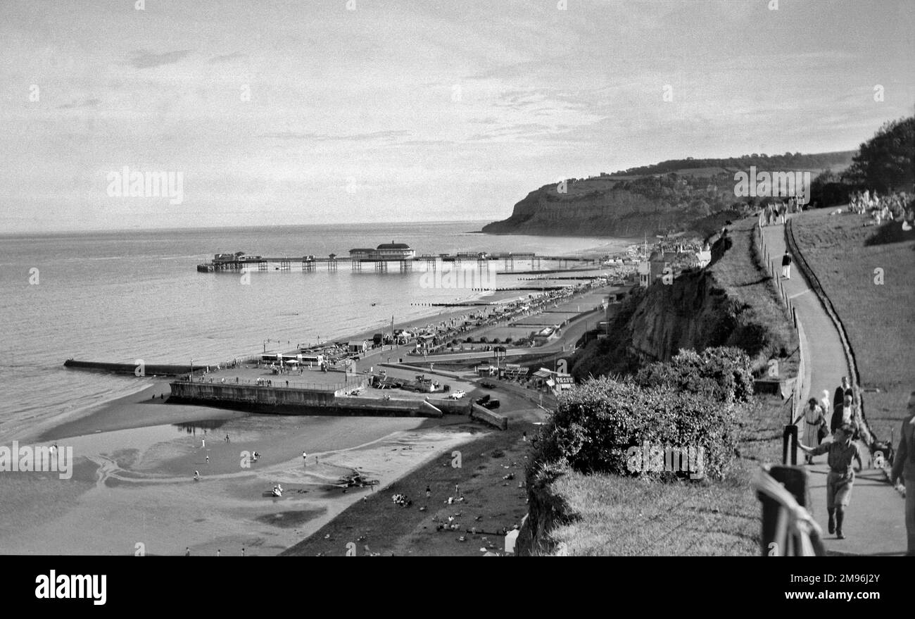 An unidentified seaside resort with a pier and a jetty, and people walking along a cliff path on the right. Stock Photo