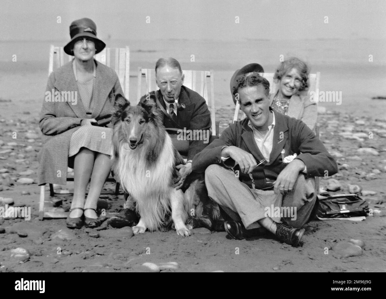 Four people (two men and two women) sitting in deckchairs on a beach with a collie dog. Stock Photo