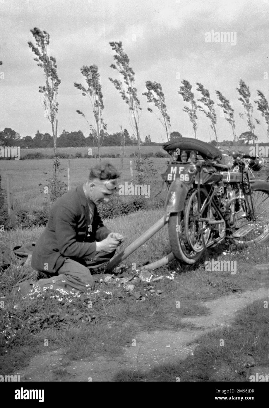 A man with his motorbike in a field.  He is either repairing the bike or picking flowers! Stock Photo