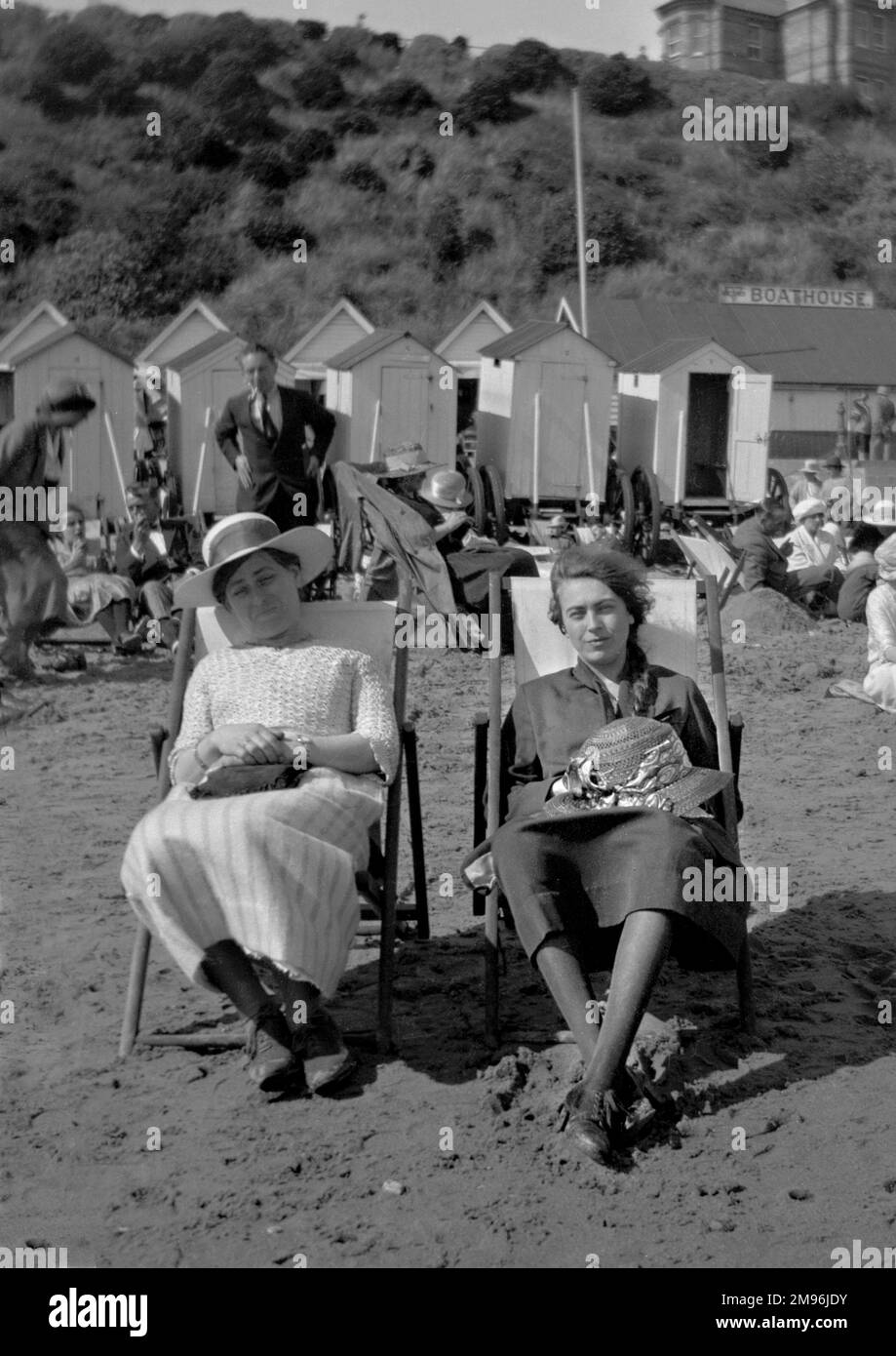 Two young women sitting on a beach in deckchairs, with bathing huts in the background. Stock Photo