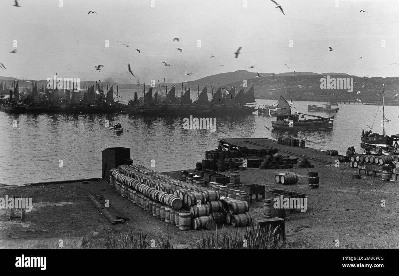 View of the docks at Barra, Outer Hebrides, Scotland, with fish barrels stacked up and boats in the harbour.  A group of fishwives can be seen on the right. Stock Photo