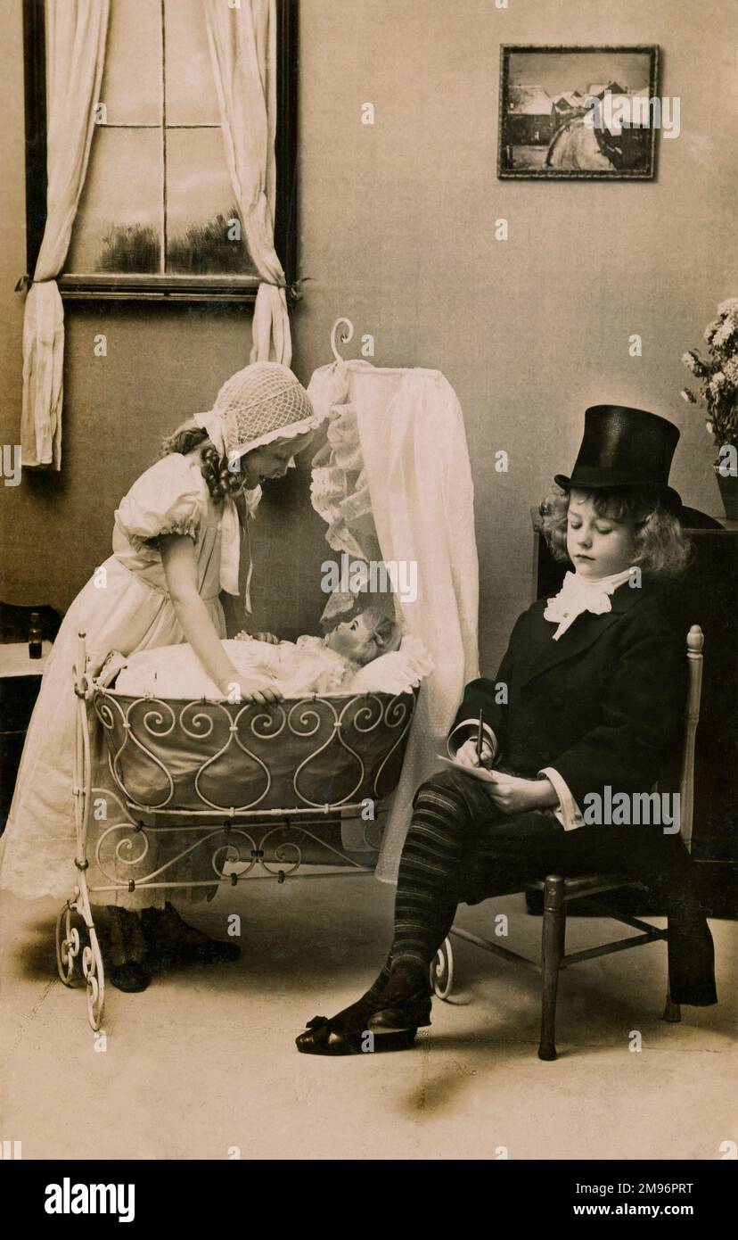 Two little children dress-up, the girl in a lace bonnet and long dress, the boy in top hat and suit, as they look after their baby doll. Stock Photo