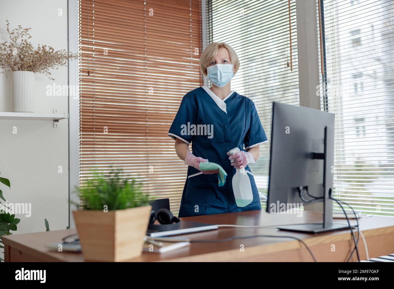 Cleaning lady in mask wiping dust off furniture in apartment. General or regular cleanup in room Stock Photo