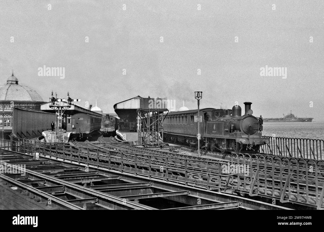 Steam engine and carriages pulling out of a coastal station, with the sea and a ship on the right. Stock Photo