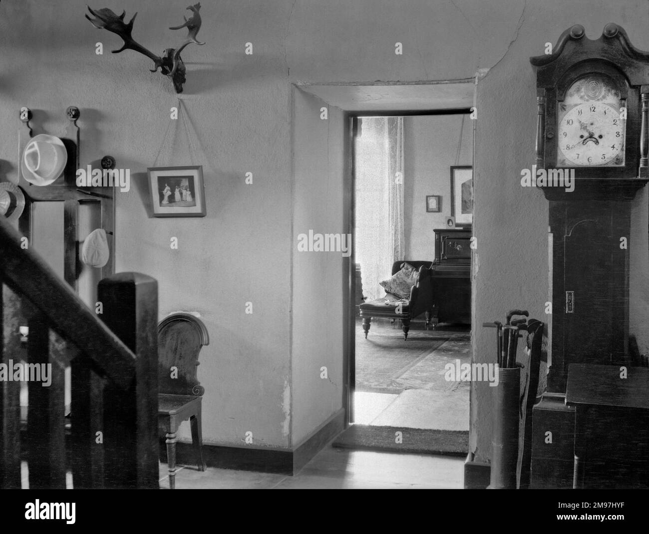 House interior showing a hallway with grandfather clock, antlers, walking sticks, hats on a stand, and another room beyond with chaise longue and piano. Stock Photo
