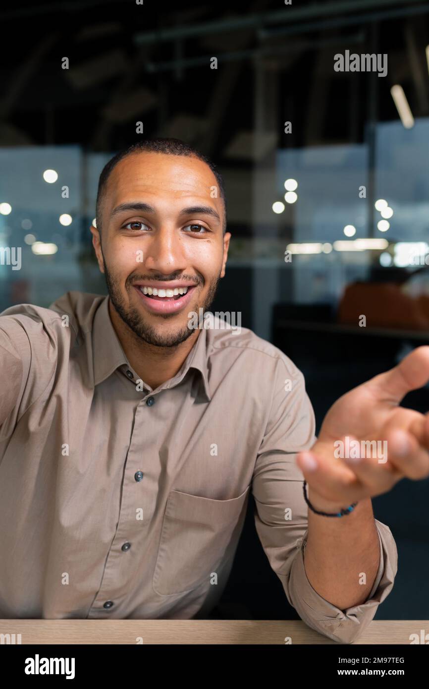 Video ringing, successful businessman looking at smartphone camera talking remotely with colleagues, Hispanic man smiling at work inside office, webcam view pov vertikal Stock Photo