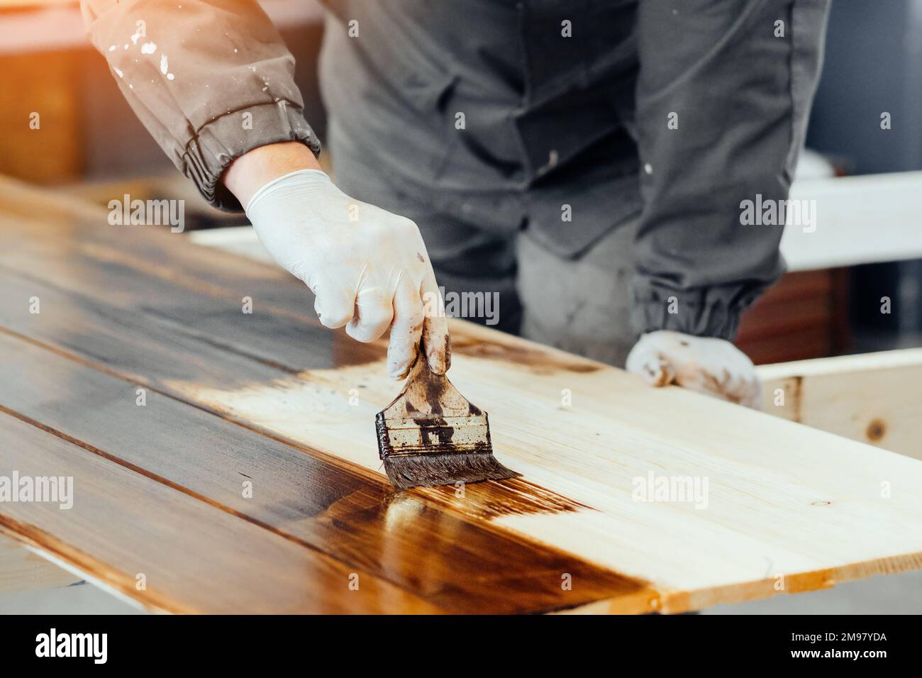 Man paints wooden boards with paint brush. Carpenter cabinetmaker varnishes wooden surface. Authentic workflow.. Stock Photo