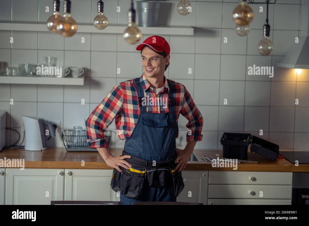 Smiling happy serviceman in the tool belt standing at the kitchen counter and looking away Stock Photo