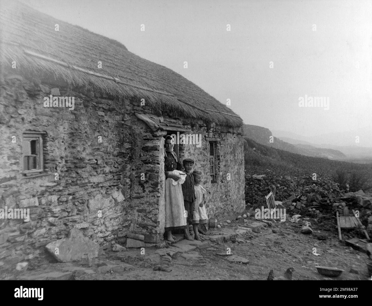 Cottage in Glen Columbkille (Glencolmcille, Gleann Cholm Cille), County Donegal, north-west Ireland.  A woman with embroidery in her hands stands at the door with two children. Stock Photo