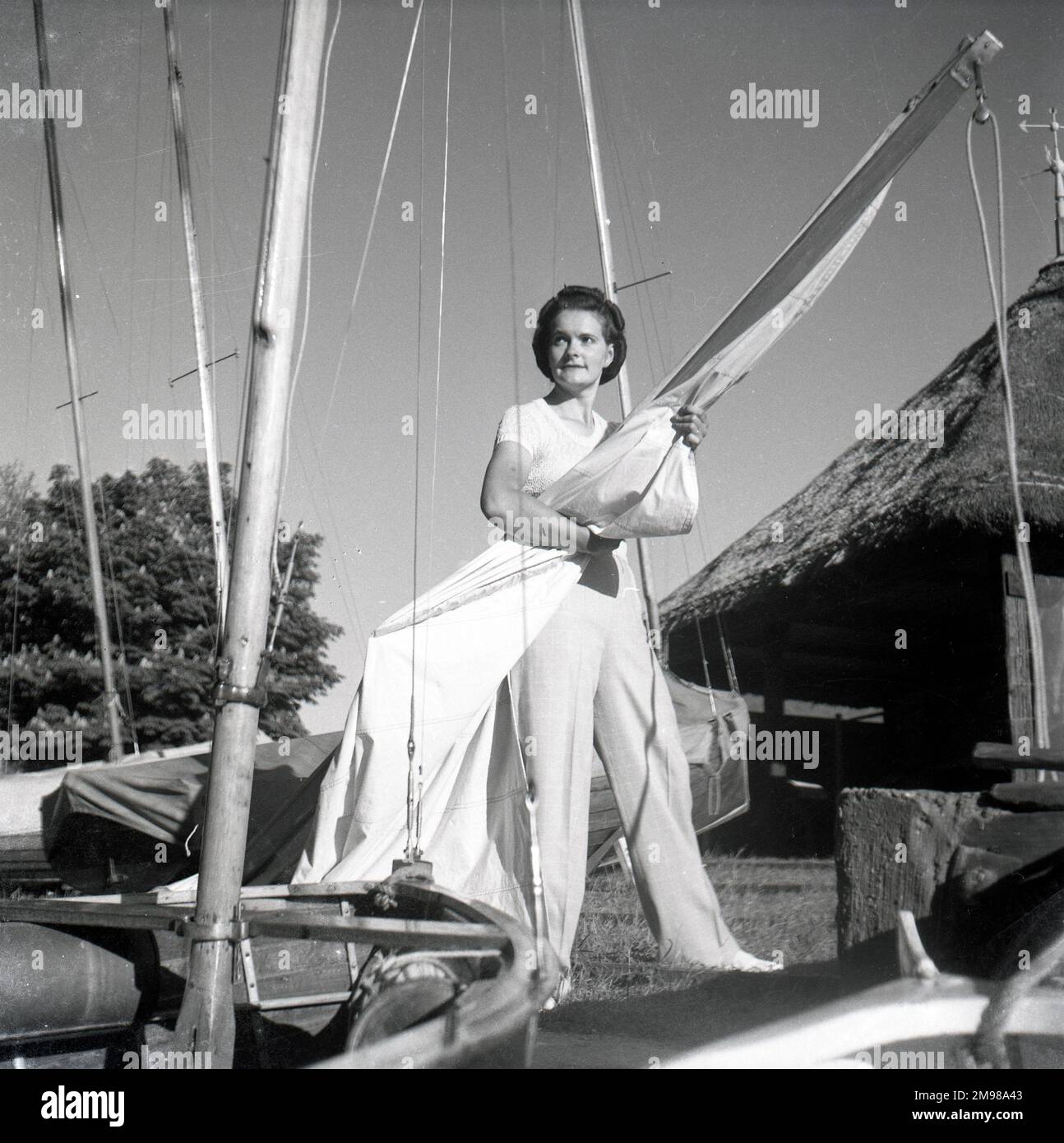 Barbara Hepworth (1903-1975), English artist and sculptor, standing alongside a yacht, with a thatched cottage on the right. Stock Photo