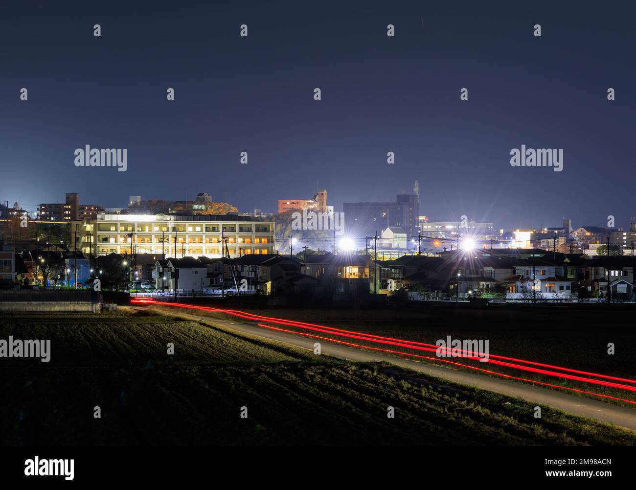 Car lights streak towards dense bright buildings on edge of town at night Stock Photo