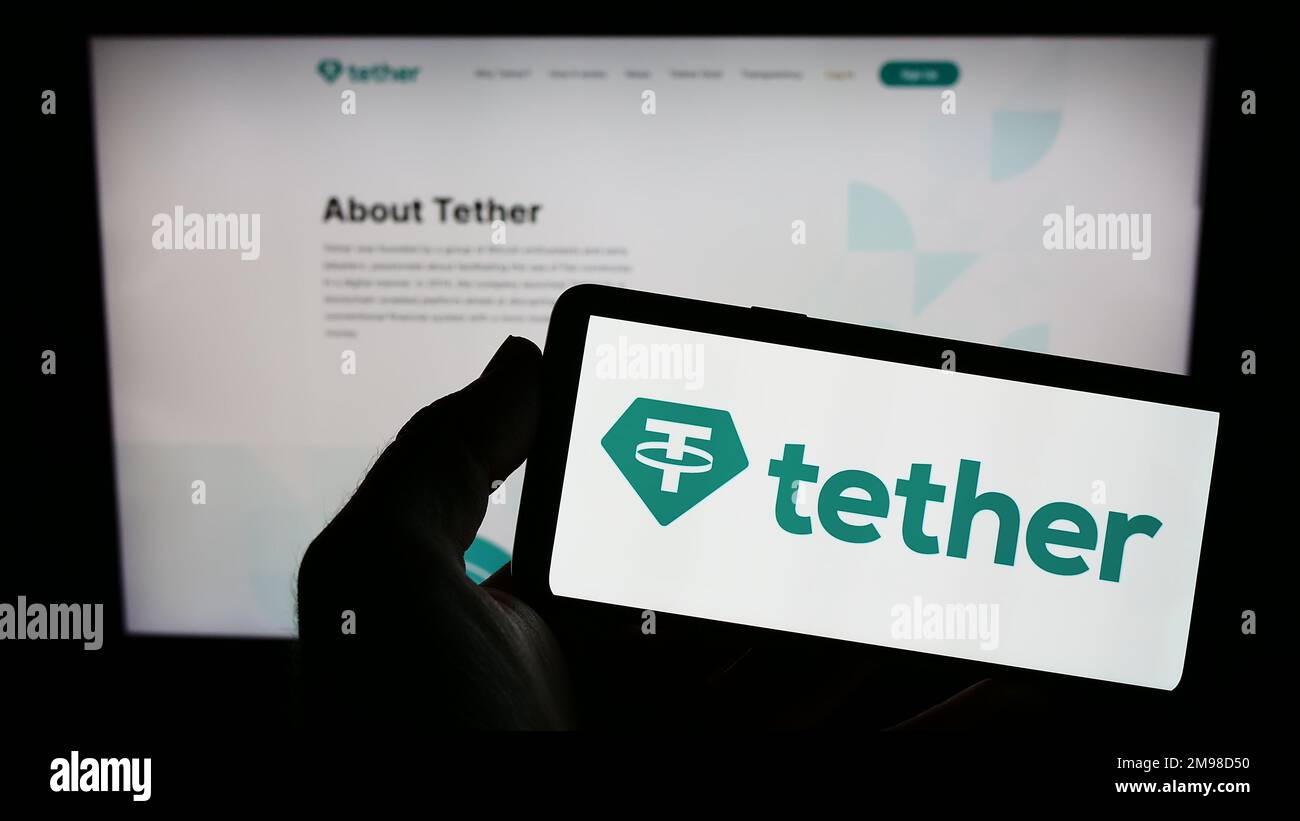 Person holding mobile phone with logo of crypto company Tether Operations Limited on screen in front of business web page. Focus on phone display. Stock Photo