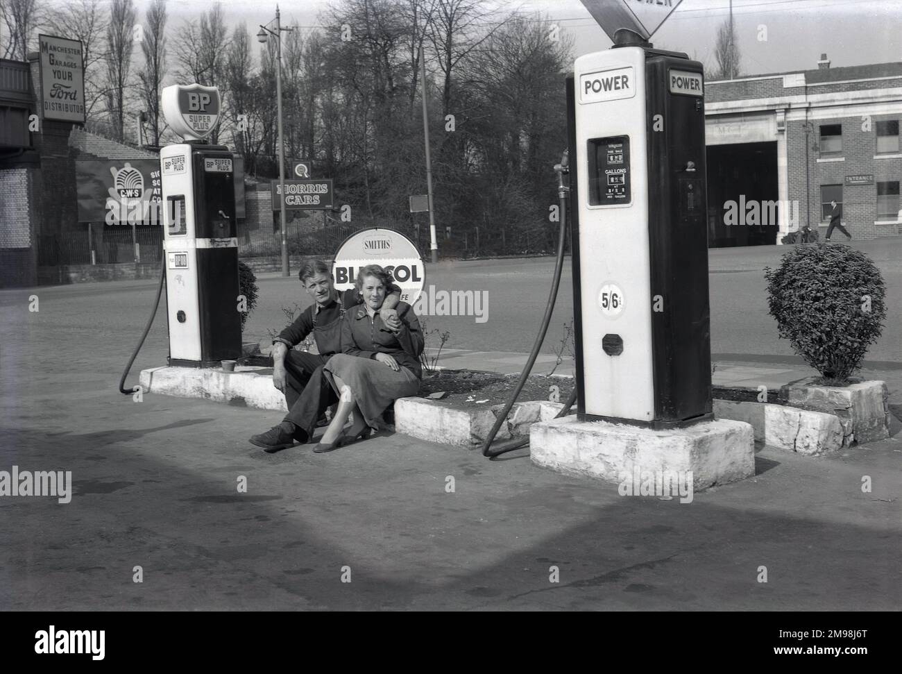 1950s, historical, a male car mechanic and female clerk of the Hargood Motor Co sitting for a photo by petrol pumps for Power and BP fuel on the forecourt of the garage, Wilmslow Rd, Parrs Wood, Didsbury, Manchester, England, UK. Price of Power petrol is 5/6d ( 5s 6d) a gallon, expensive, as it was the time of the Suez Crisis. A large bus depot servicing the city of Manchester is across the road. A sign for Morris Cars can be seen at the entrance to the railway bridge, with the East Didsbury & Parrs Wood railway station nearby. Stock Photo