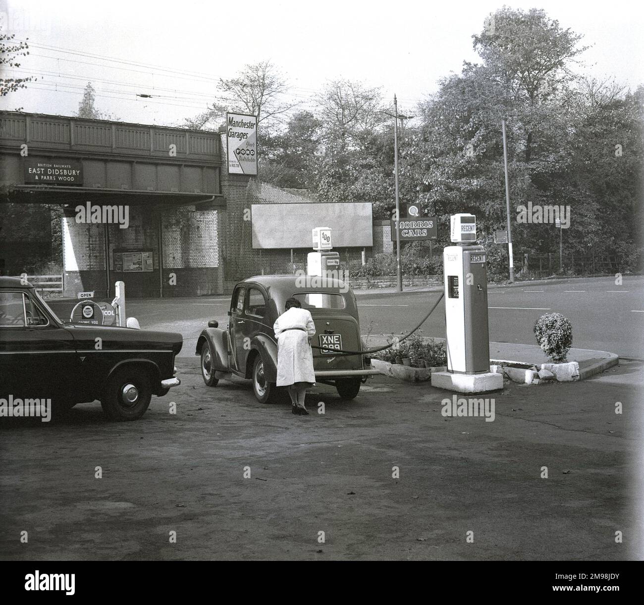 Late 1950s, historical, a female garge attendant putting petrol in a Ford Popular car at the Hargood Motor Co on the Wilmslow Rd, Parrs Wood, Manchester, England, UK. The fuel is Regent, a brand of The Regent Oil Company, set up in the UK in 1947 by Texaco of America and Trindad Leaseholds. Behind the garage, a railway bridge, sign for station, saying 'British Railways', East Didbsbury & Parrs Wood', Stock Photo