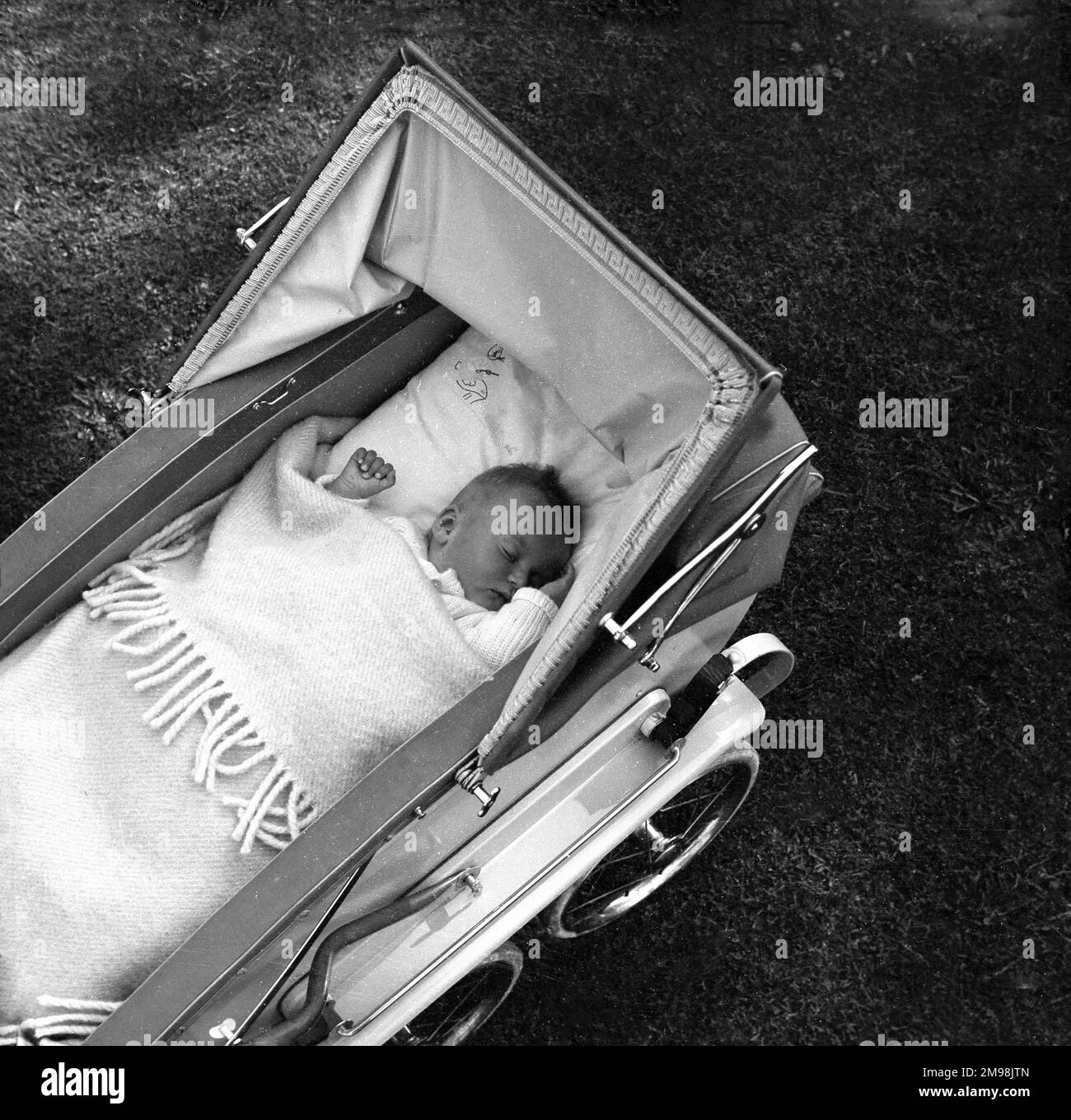 1950s, historical, view from above of a baby lying on its back, asleep, under a blanket, in a traditional four-wheeled coach-built pram or baby carriage, England, UK. This type of baby transport provided a comfortable, enclosed space for the small child to rest and sleep in and they would be facing the parent when pushed, enabling eye contact and interaction when awake. Stock Photo