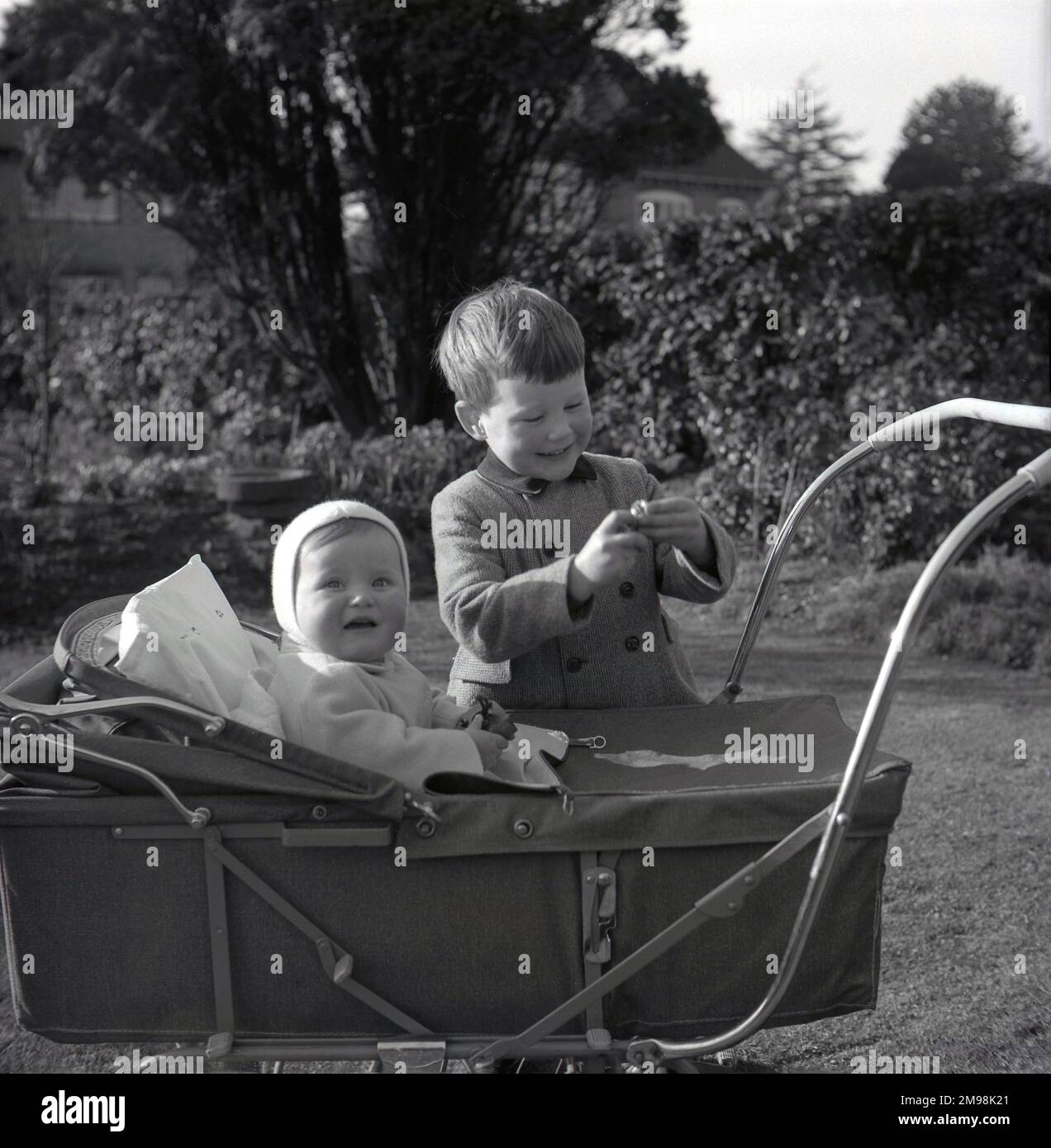 1960s, historical, outside in a garden, a young boy standing beside his infant brother who, with a cushion behind him, is sitting up in a metal framed pram of the era, England, UK. This type of baby or infant carring carriage was the same design of the traditional four-wheeled coach-built pram, where the child is facing the person pushing the pram, but was a lighter and smaller version, having a frame which meant that that it folded down when not in use. Stock Photo