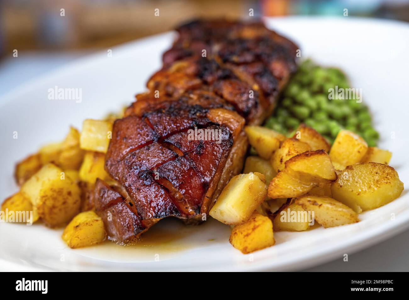 Baked fried pork belly with crispy skin and potato and pea on white plate on table, closeup. Stock Photo