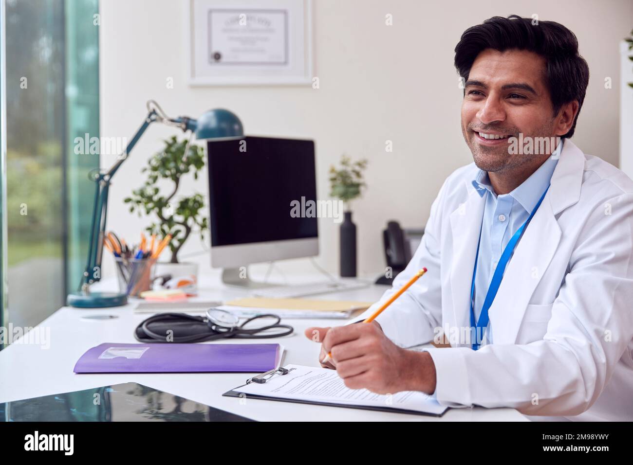 Male Doctor Or GP Wearing White Coat Sitting At Desk In Office Writing Notes On Clipboard Stock Photo
