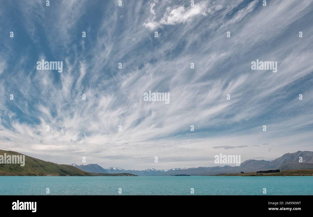 Wispy clouds and blue skies over the turquoise blue water of Lake Tekapo in New Zealand with snow capped mountains in the distance Stock Photo