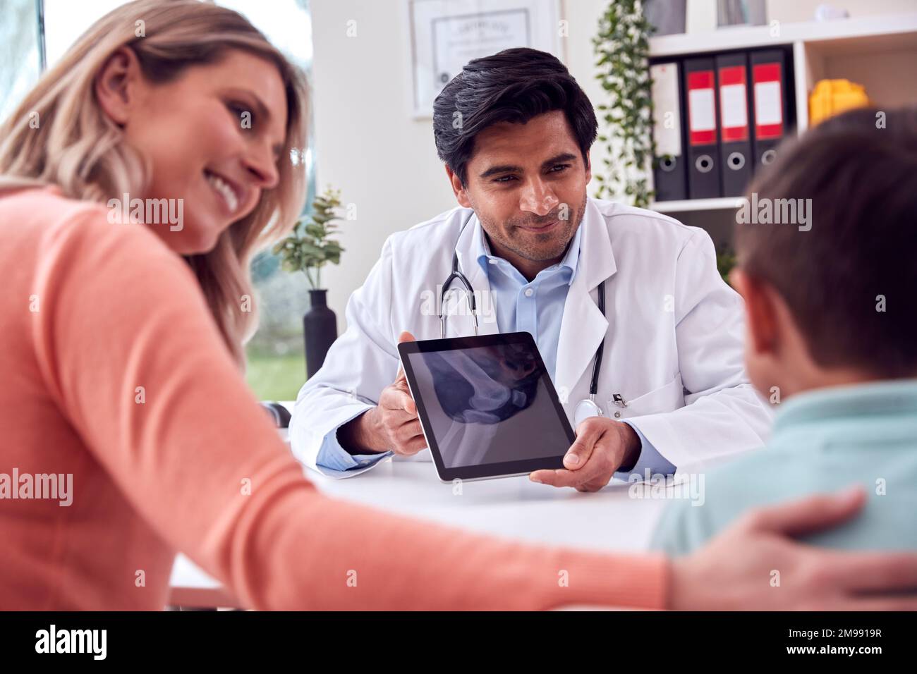 Doctor Or GP In White Coat Meeting Mother And Son For Appointment Looking At Scan On Digital Tablet Stock Photo