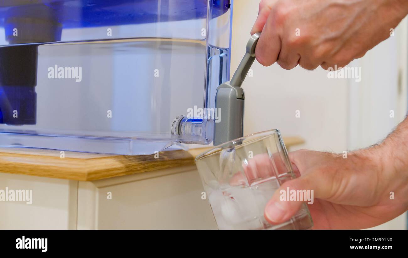 Dispensing fresh filtered water from home water purification pitcher. Man pouring clean water into a glass with ice from container water spout. Stock Photo