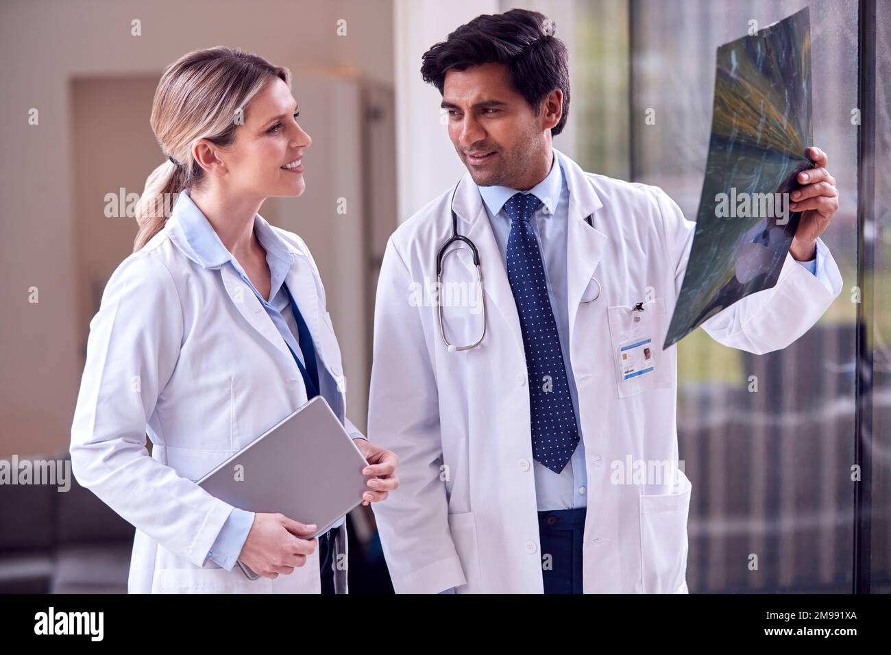 Male And Female Doctors Wearing White Coats Standing In Hospital Corridor Looking At CT Or MRI