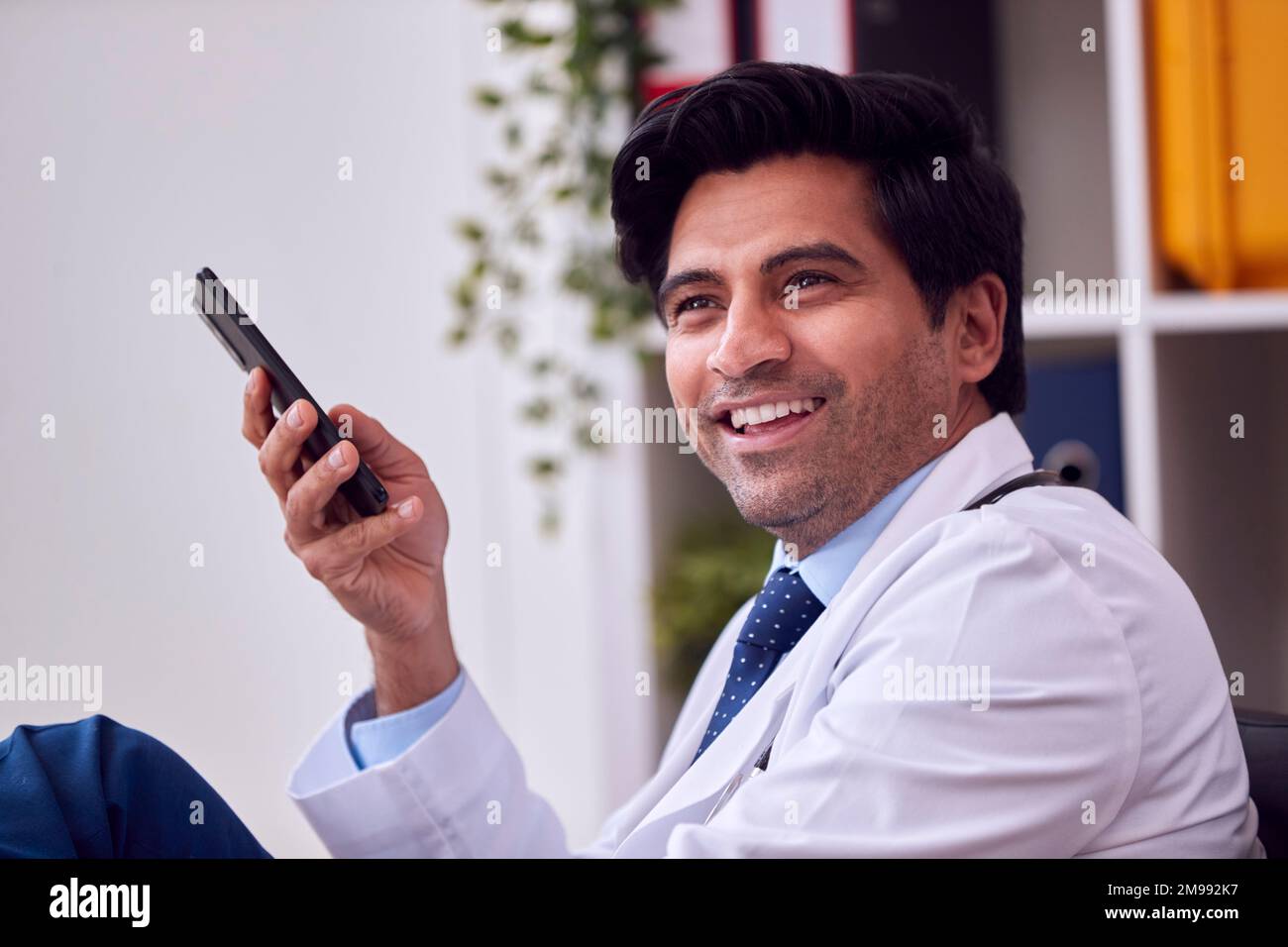 Smiling Male Doctor Or GP Wearing White Coat Sitting At Desk In Office With Mobile Phone Stock Photo
