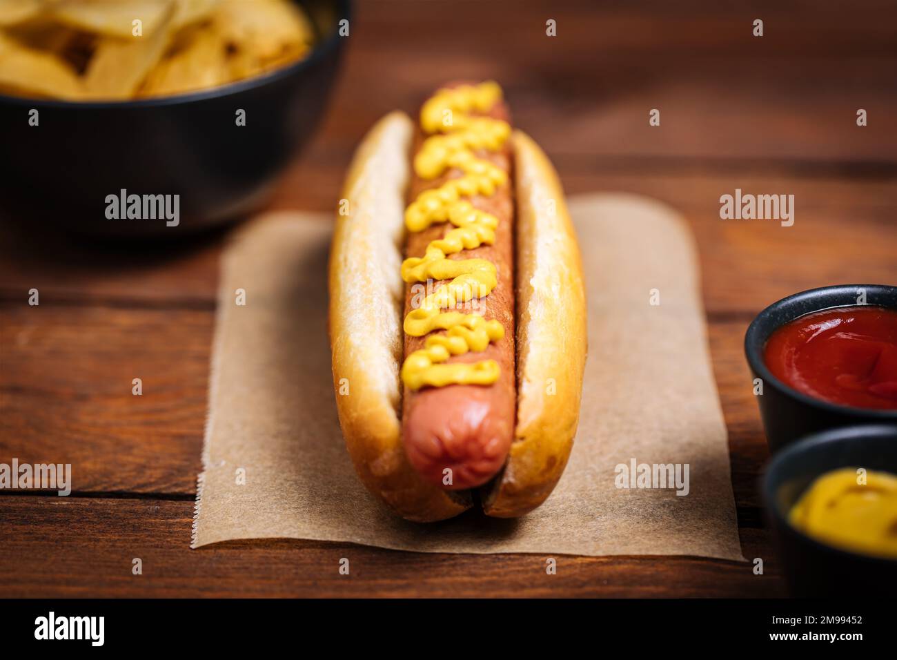 Classic fast food hot dog served with mustard on a rustic wooden board. Served with potato chips and sauces. Stock Photo