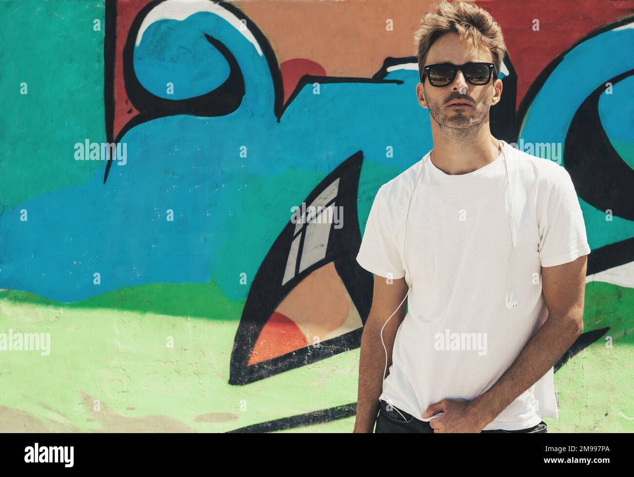 Young bearded handsome man standing next to a colorful wall background wearing an empty white t-shirt and sunglasses. Horizontal mock up style. Stock Photo