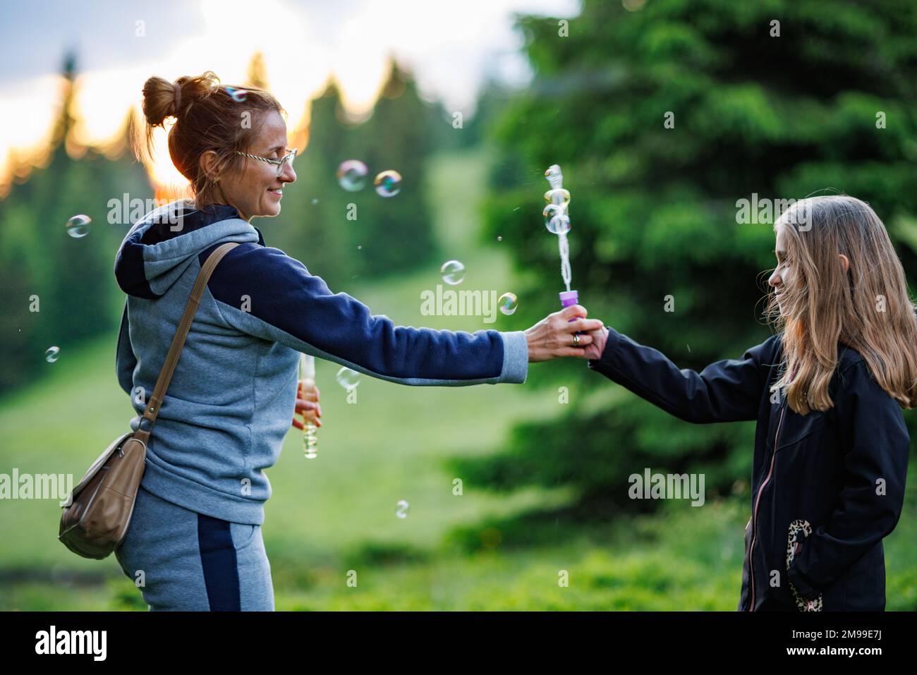 Happy kind caring mother and her cute smiling cheerful daughter with loose hair blow bright soap bubbles of different sizes, spring spruce green fores Stock Photo