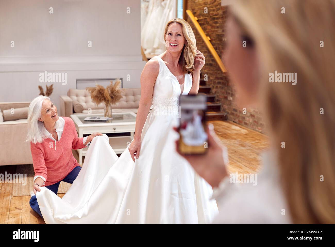 Grandmother With Adult Daughter And Granddaughter Trying On Wedding Dress Taking Picture On Phone Stock Photo