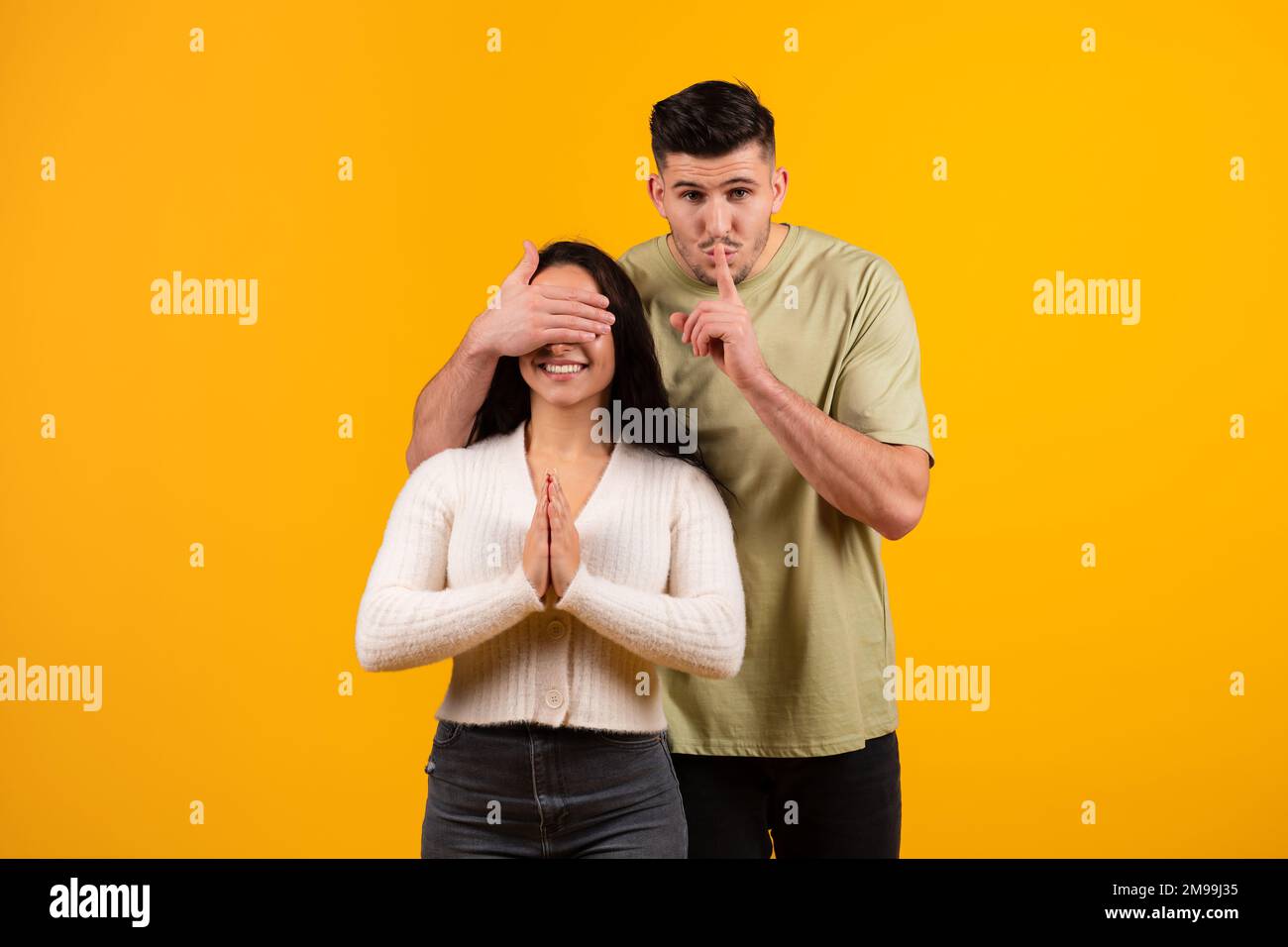 Cheerful millennial arabic guy closes eyes to amazed wife, makes shhh sign, preparing surprise for anniversary Stock Photo