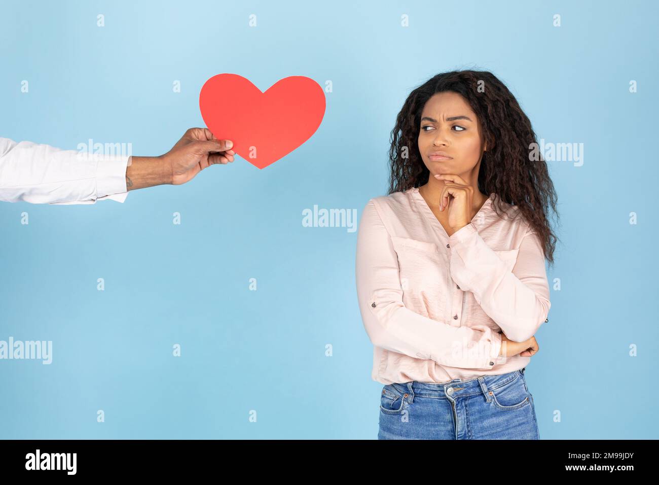 Yes or no. Man giving paper red heart to young woman, lady thinking about date invitation answer, blue studio background Stock Photo