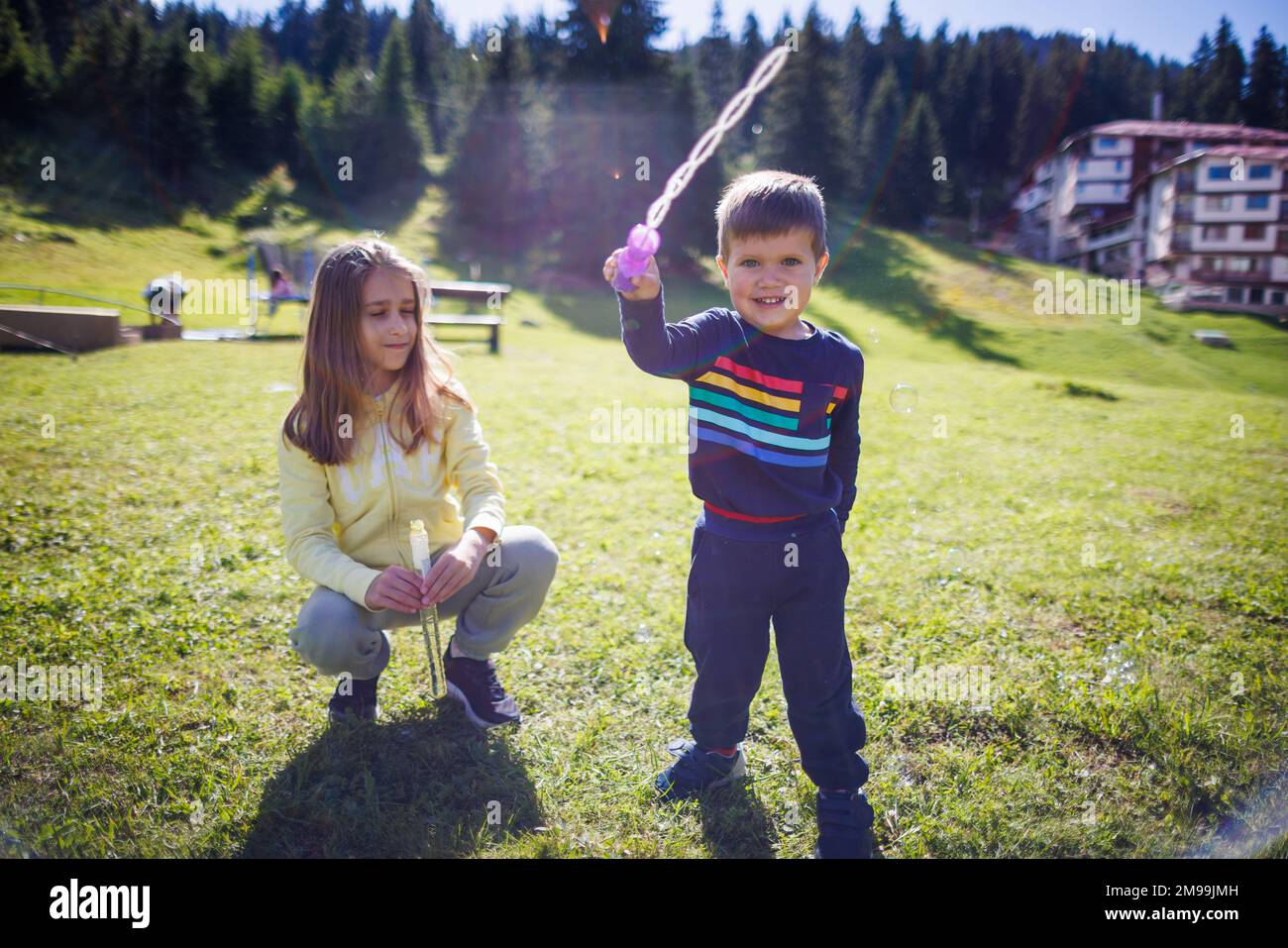 An older sister with long hair in a yellow tracksuit plays with her younger brother in a blue suit and teaches him how to blow soap bubbles on a green Stock Photo