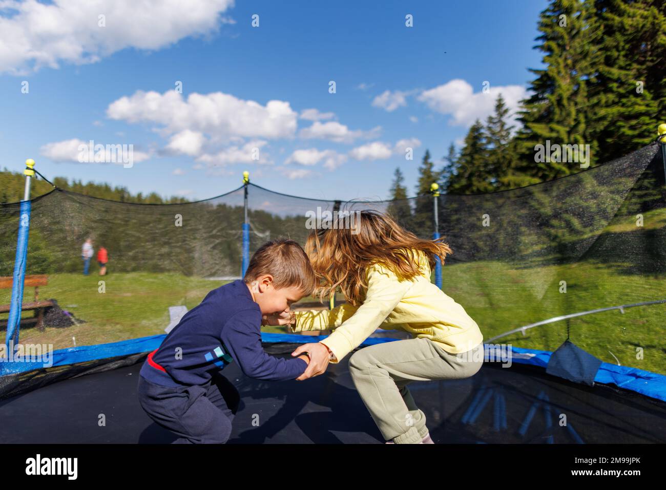 An older sister with flowing long hair in a yellow tracksuit and her little brother in a cute blue suit are jumping together on a netted trampoline in Stock Photo