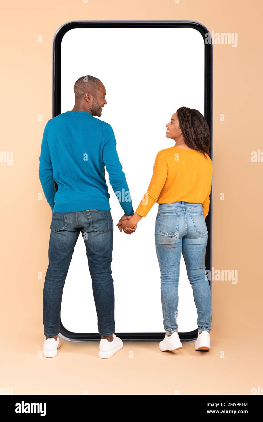 Happy black couple standing near huge phone with empty screen, standing back to camera, holding hands and smiling Stock Photo
