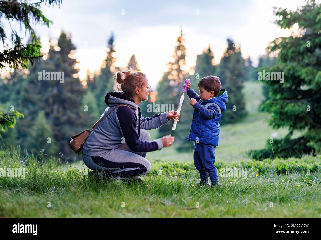 A happy smiling caring mother in a warm gray sports suit plays with her little cheerful son in a cute blue suit on a green lawn and teaches him to blo Stock Photo