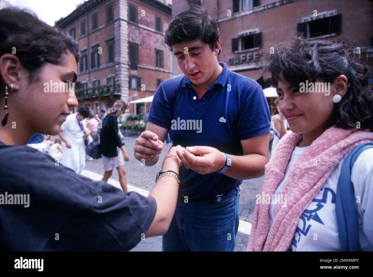 A young Italian man selling wrist bracelets ties one on a young woman tourist in a Rome piazza. Stock Photo