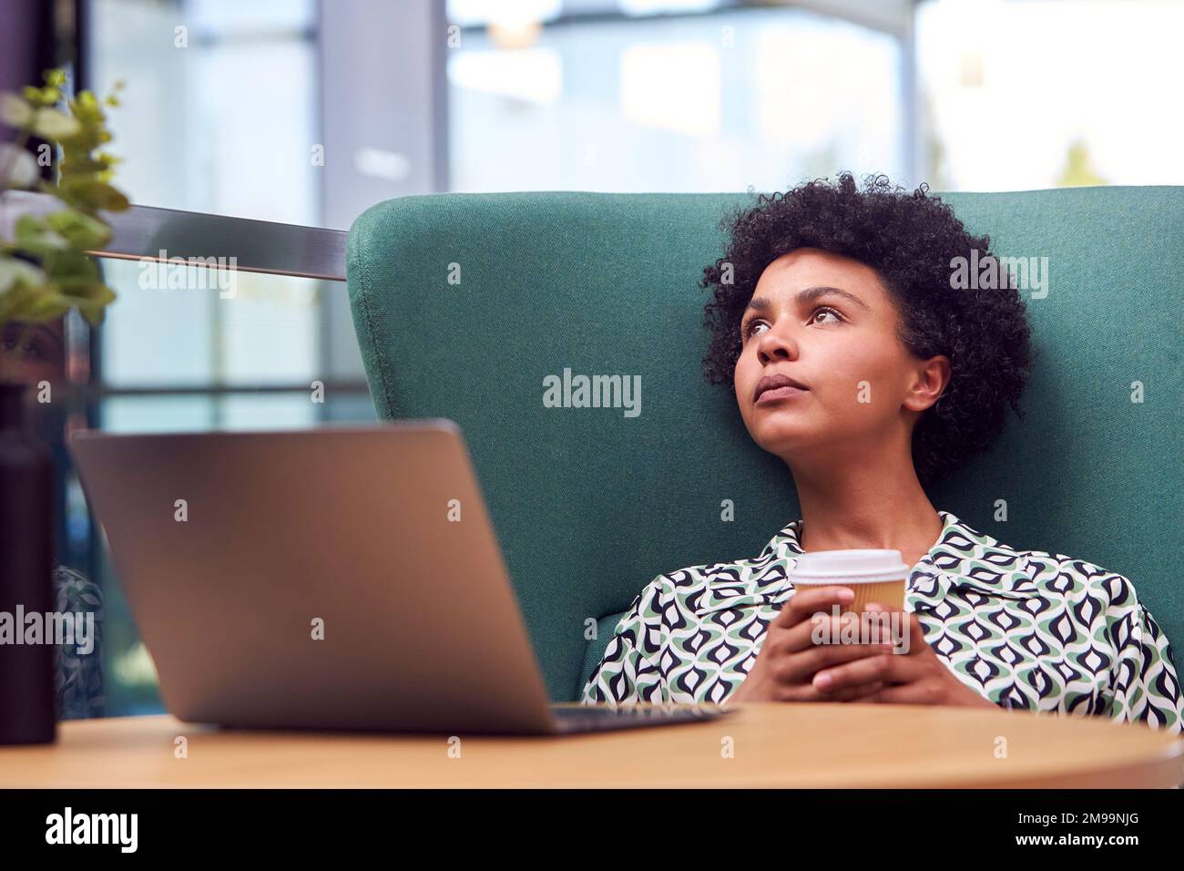 Thinking Businesswoman Using Laptop Working At Table In Breakout Seating Area Of Office Building Stock Photo