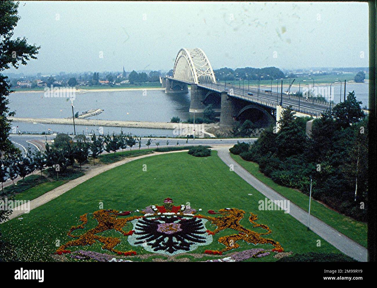During Operation Market Garden in September 1944, the British and Americans, with Polish support, launched a joint air and land assault northwards from Belgium and then via Holland, in an attempt to outflank German defences along the River Rhine. The bridge here at Nijmegen was the target of the American 82nd Airborne Division, which was tasked to capture it so that the advancing tanks of the British Grenadiers could cross the river and move on towards Arnhem. The Coat of Arms in the foreground is that of Nijmegen, and the grassed area is bordered with Canadian Sugar Maple trees presented by Stock Photo