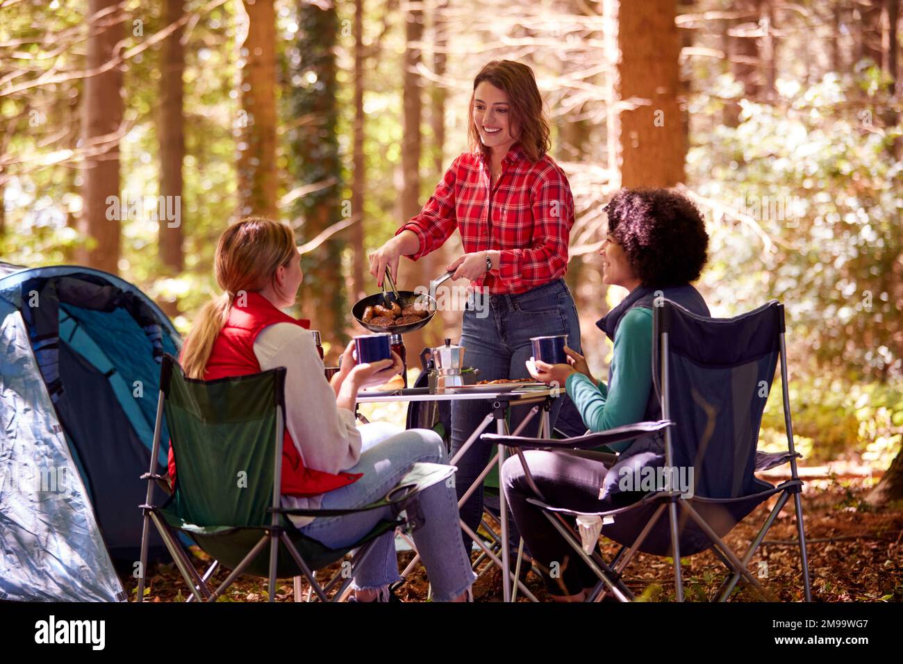 Group Of Female Friends On Camping Holiday In Forest Eating Meal Sitting By Tent Together Stock Photo