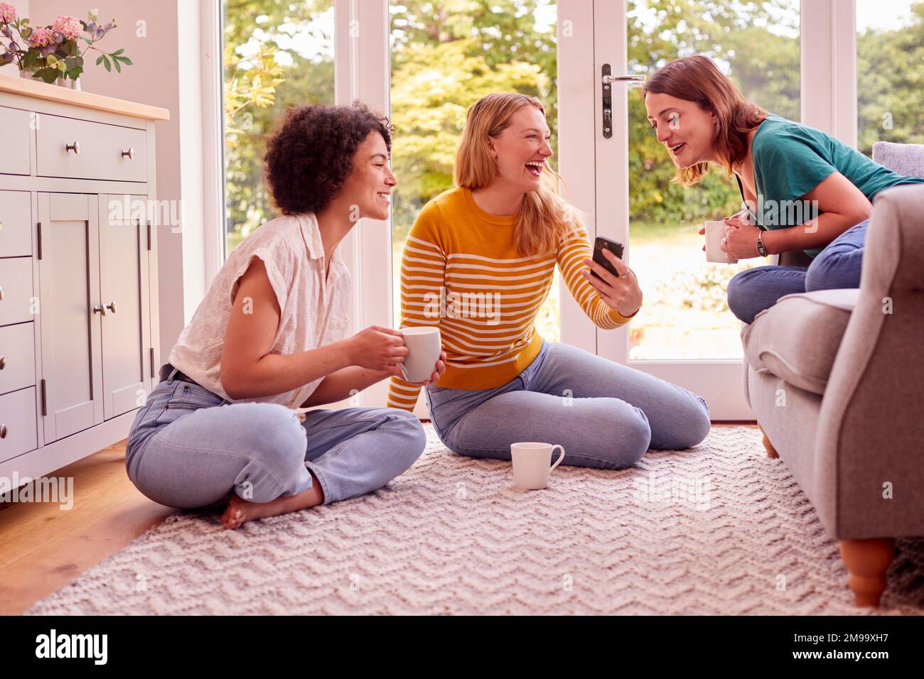 Group Of Female Friends Relaxing At On Sofa At Home Looking At Mobile Phone And Drinking Coffee Stock Photo