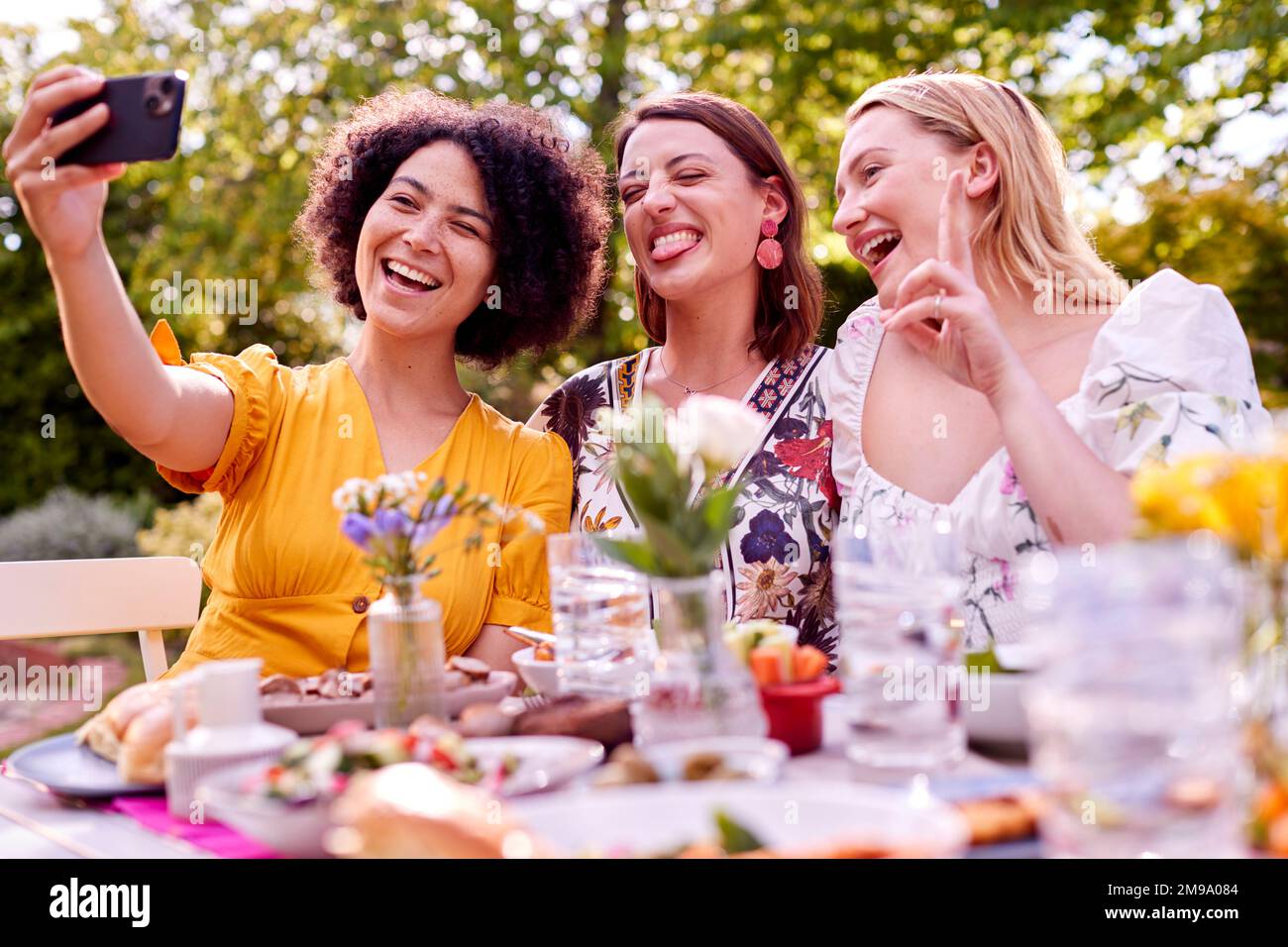 Three Female Friends Taking Selfie On Mobile Phone Eating Meal Outdoors In Summer Garden At Home Stock Photo