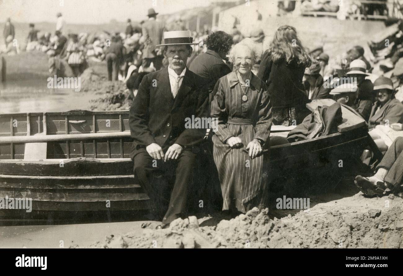 An elderly couple pose for a photograph sitting on the side of a rowing boat on the beach at a busy British seaside resort. Note the lapel cord on the gentleman's fine boater hat! Stock Photo