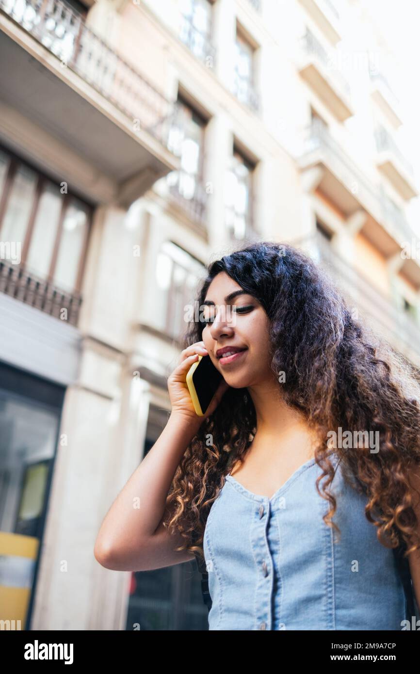 Low angle of glad Hispanic indian woman with backpack answering phone call and looking away. While walking on historic city street in daytime Stock Photo