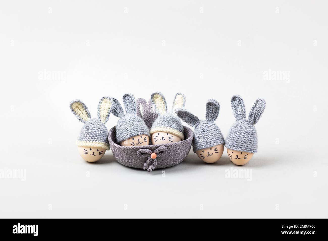 Five Easter eggs with funny faces and gray crocheted hats with bunny ears in a decorative basket on a gray background. Happy Easter concept. Greeting Stock Photo