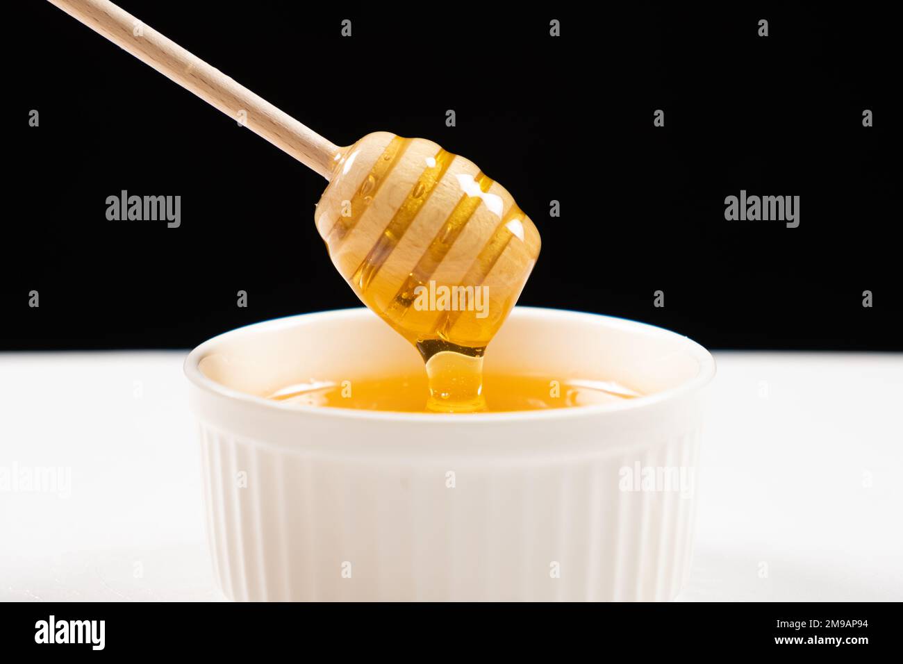 Honey in a white ceramic bowl with a wooden spoon on a black background. Stock Photo
