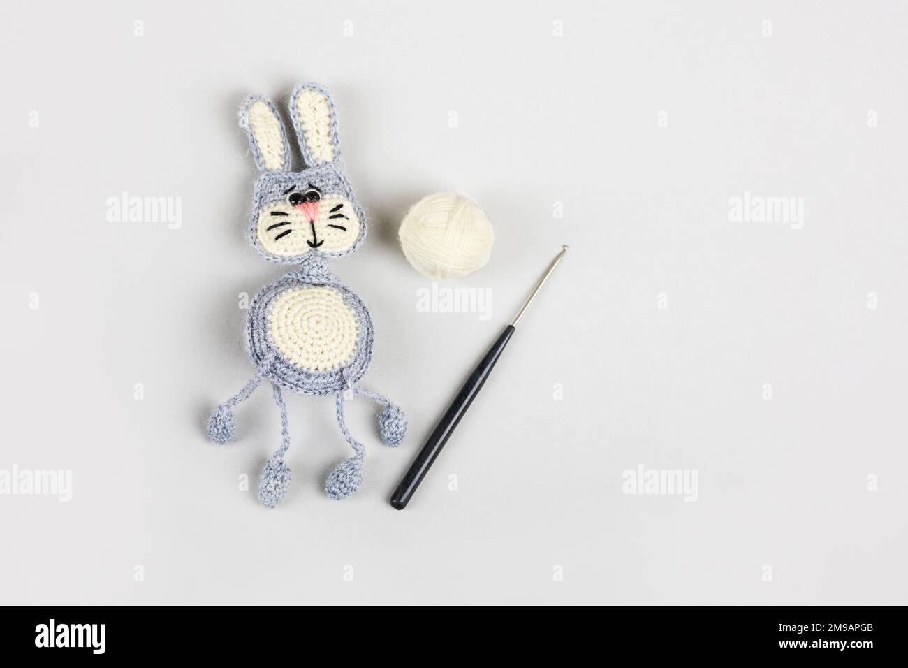Knitted children's toy hare or bunny with a balls of thread and a crochet hook on a gray background. Stock Photo