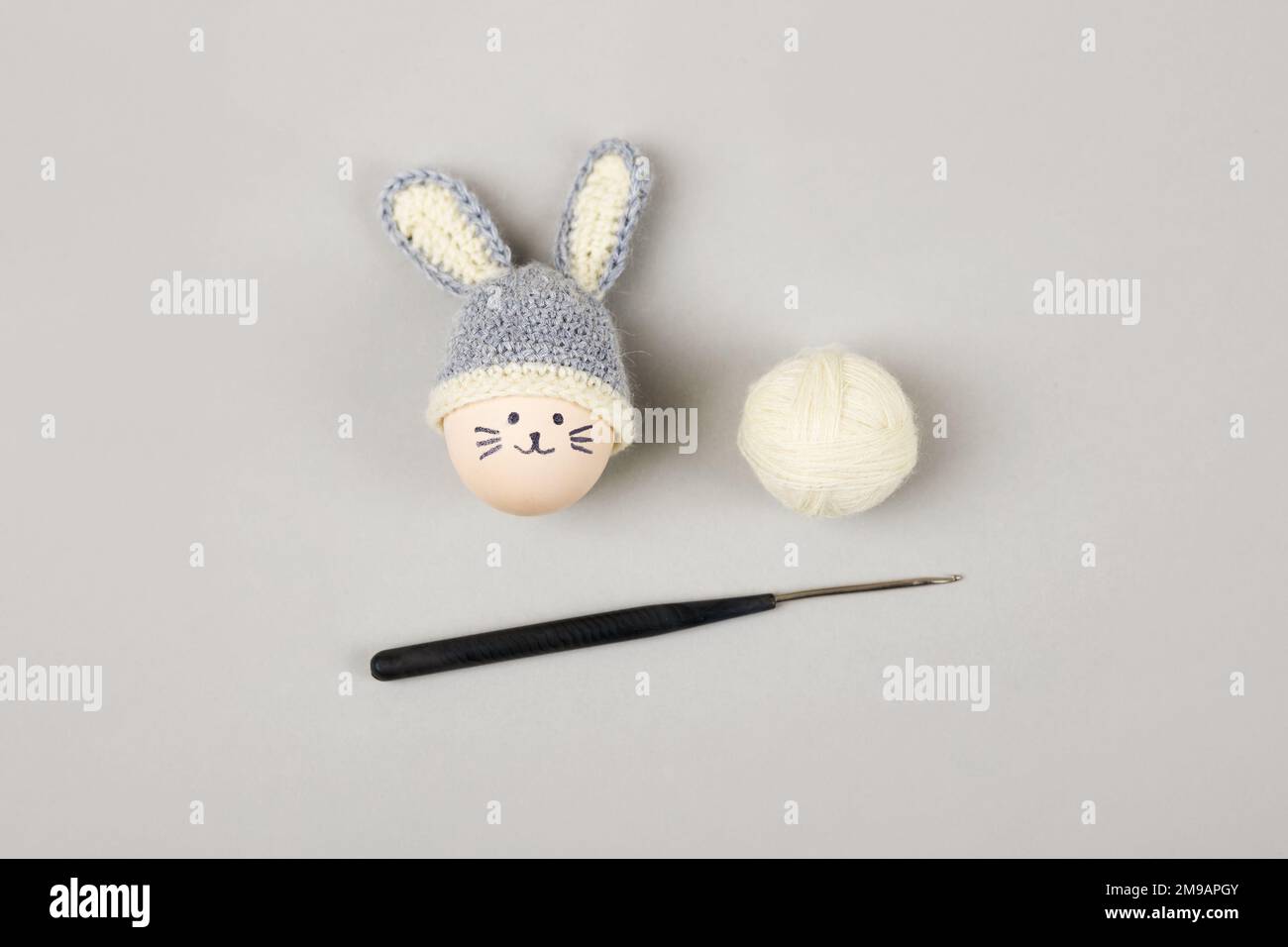 Easter egg with funny face and gray crocheted hat with bunny ears with a balls of thread and a crochet hook on a gray background. Stock Photo